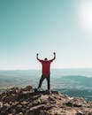 一名男子在晴朗的天空下，站在岩石山顶上，俯瞰广阔的风景，庆祝胜利