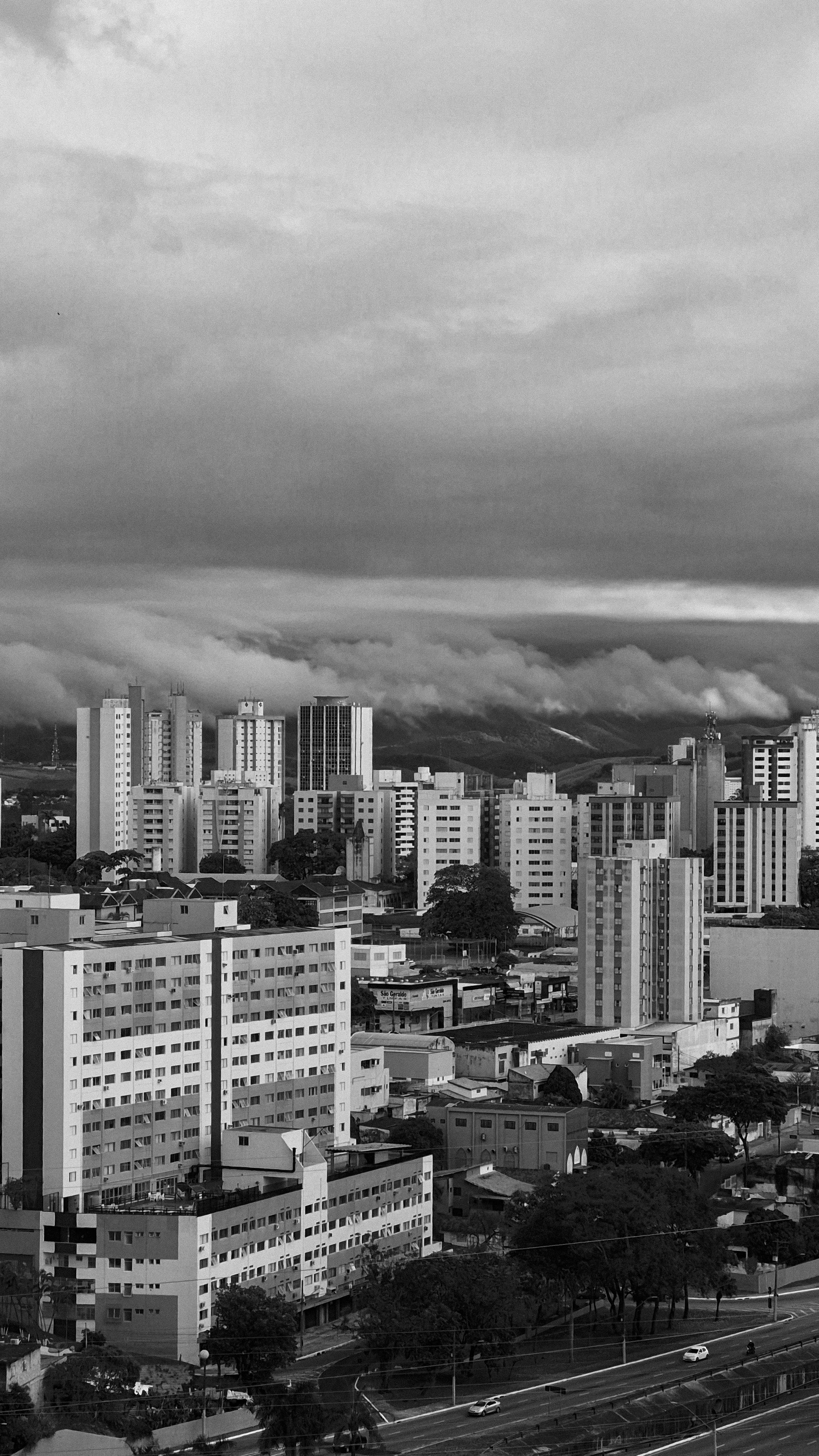 Black and white aerial view of a modern city skyline under a dramatic cloudy sky.