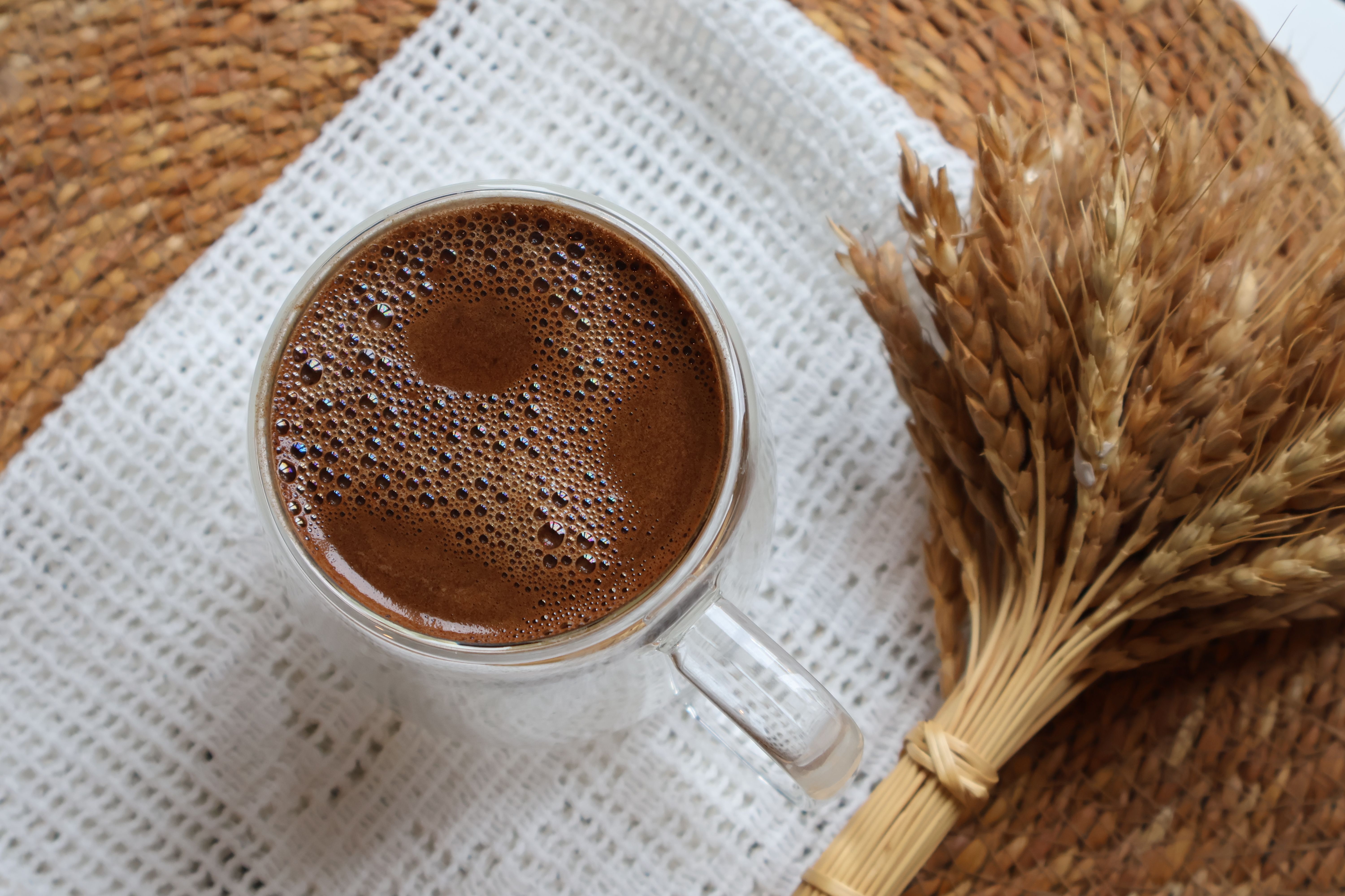 A steaming cup of coffee with foam alongside a wheat bundle on a rustic woven mat.