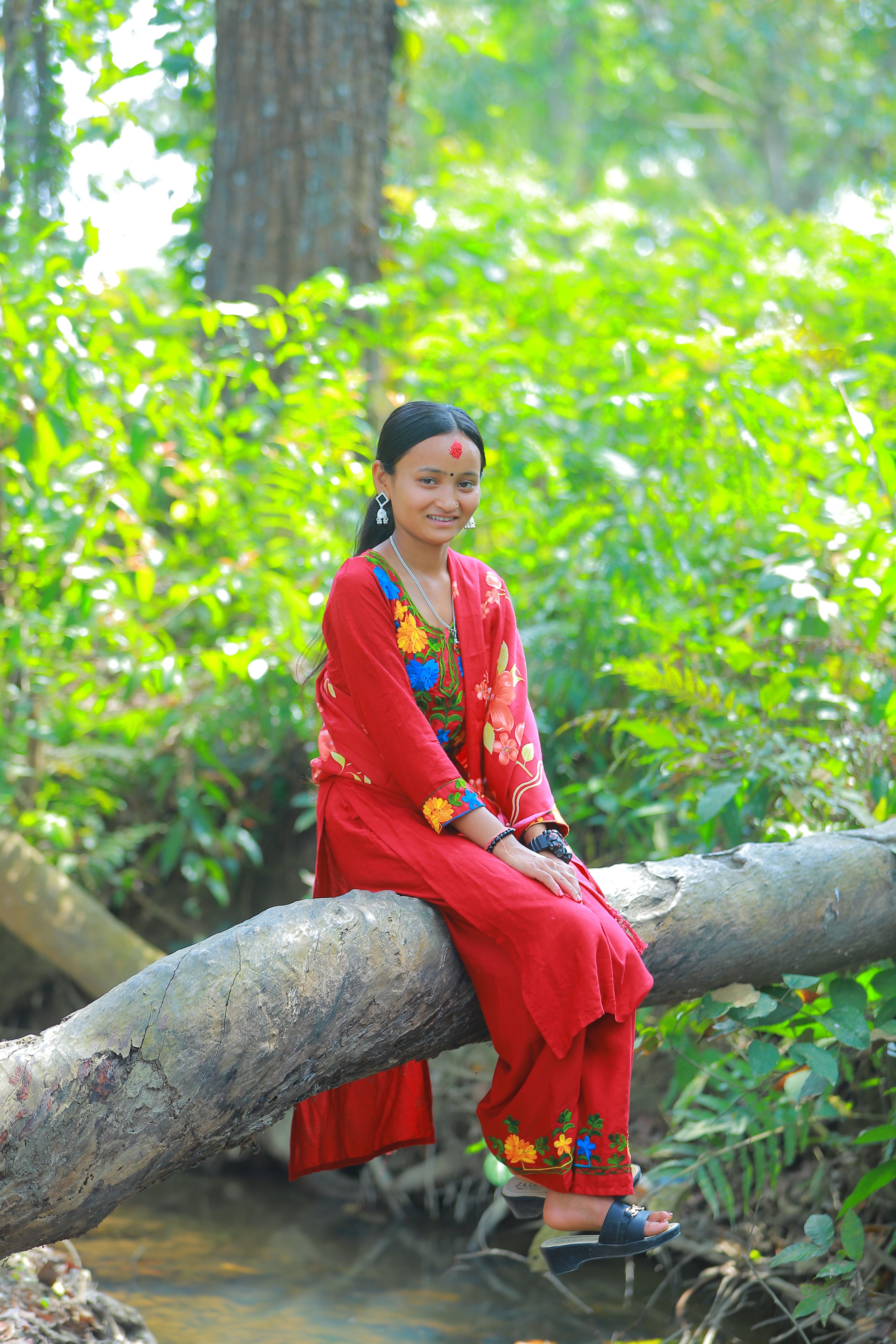 A woman in traditional attire sits boldly on a tree log in a lush, green forest.