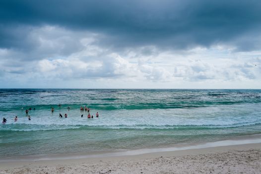 Beachgoers enjoy ocean waves under a dramatic cloudy sky.
