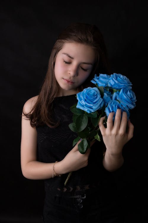 Gentle girl in black dress with bunch of fresh blue roses standing on black background
