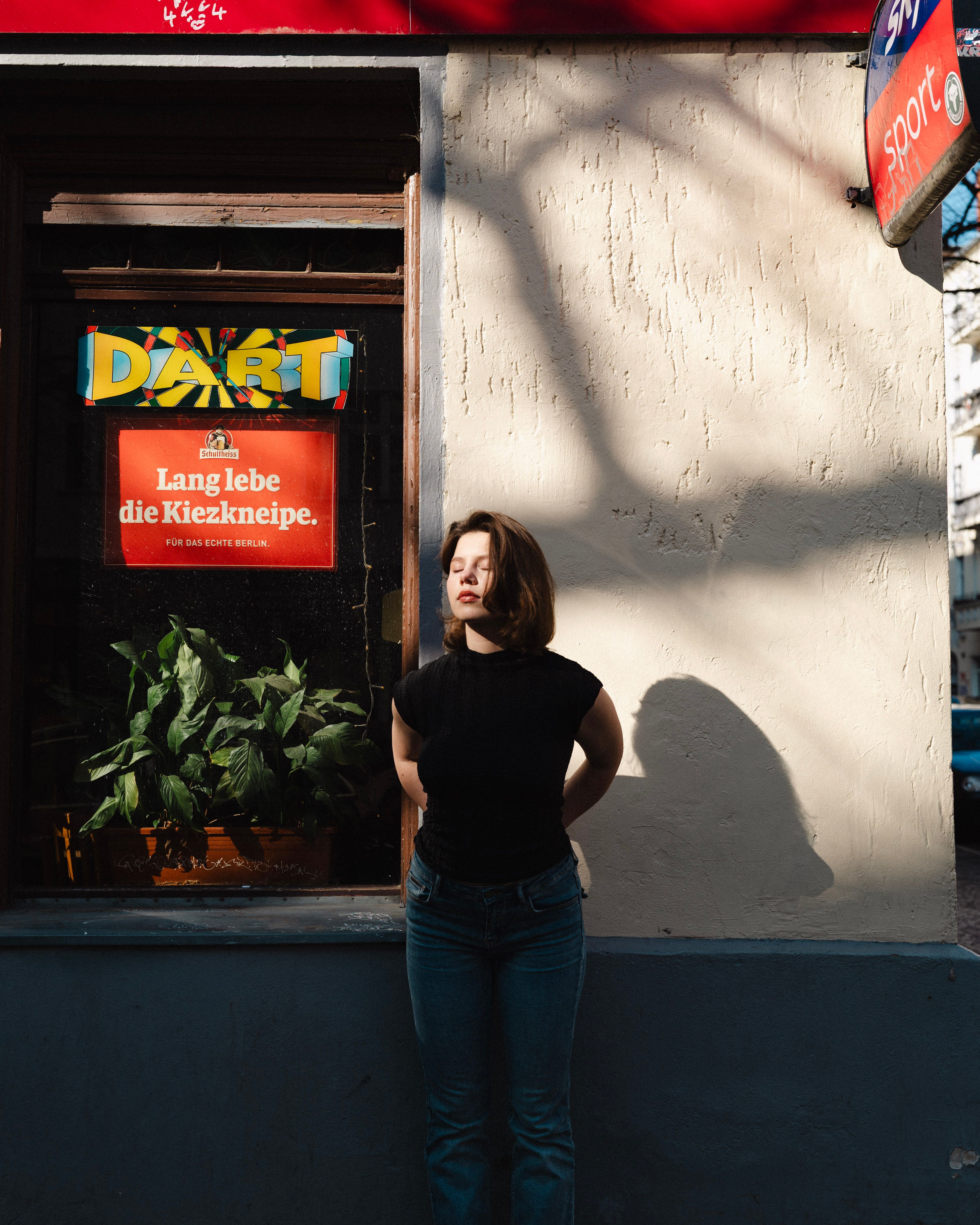 A woman stands in front of a wall displaying a colorful Dart sign and plants, under bright daylight in Berlin.
