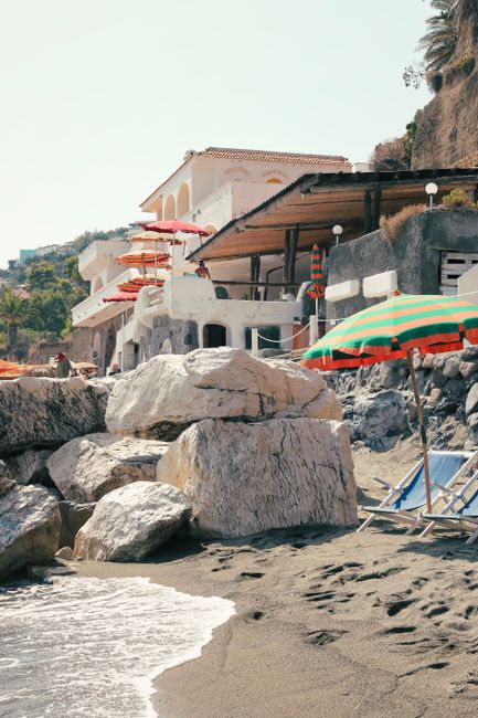 Coastal view showcasing a beach with colorful umbrellas, lounge chairs, and rocky shoreline under clear skies.