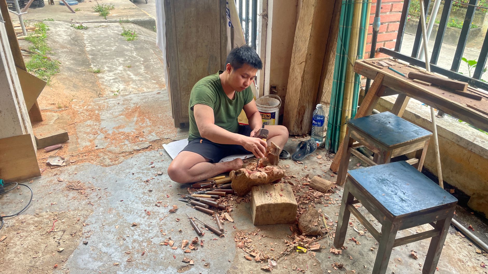 A craftsman intricately carves wood indoors, surrounded by tools and wood shavings.