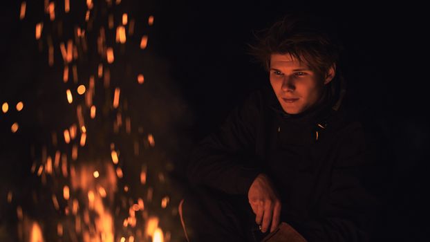 A young adult sits pensively by a campfire, with sparks illuminating the night.