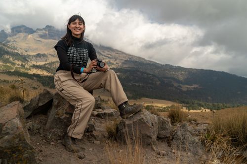 Free A smiling photographer relaxes on rocks with her camera, surrounded by majestic mountains. Stock Photo