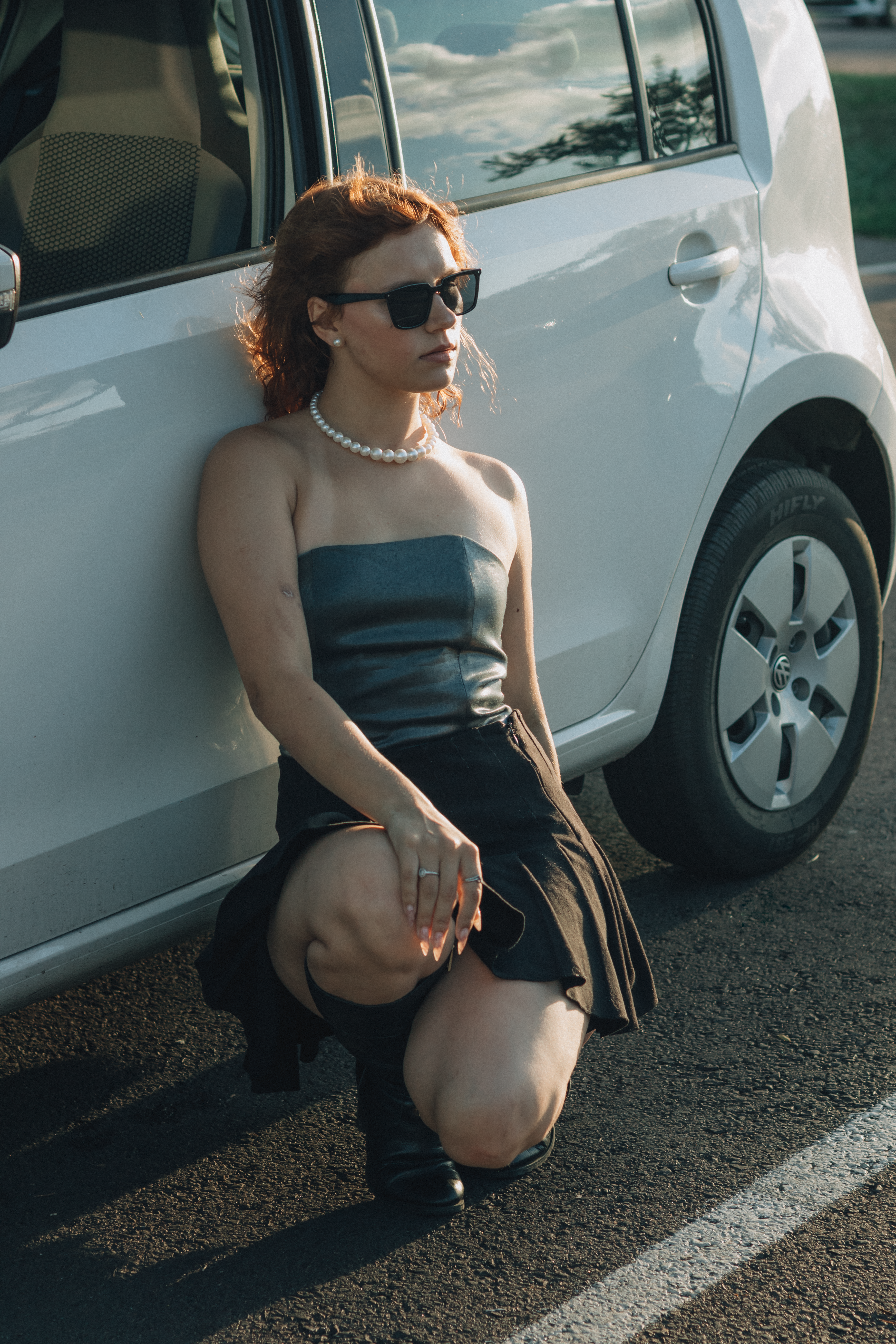 Stylish woman kneeling by car, wearing sunglasses and a pearl necklace.