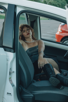 Woman in boots and dress seated in car, serene expression, parking lot background.