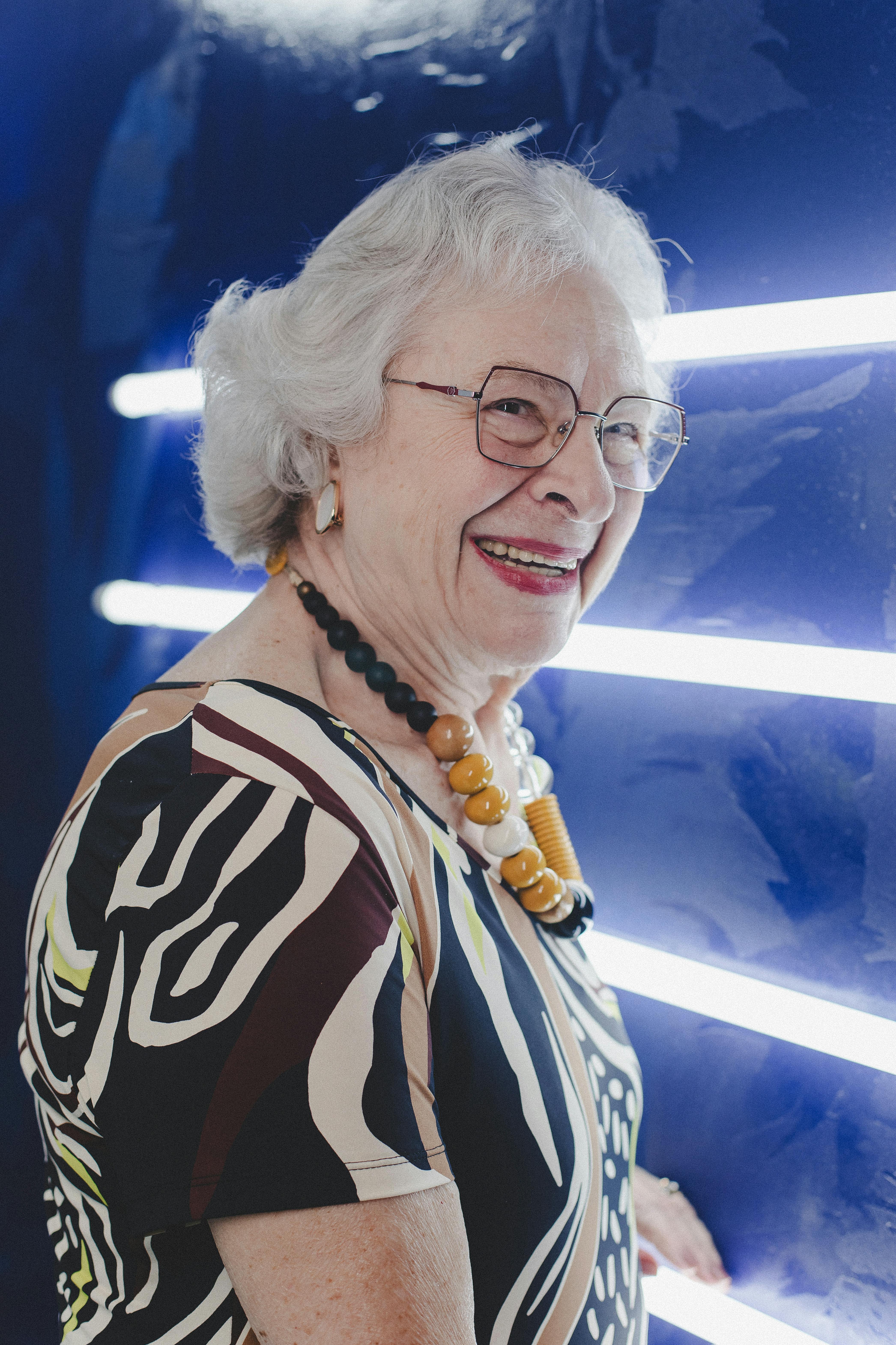 Elderly woman with white hair and glasses smiling in front of bright neon lights.