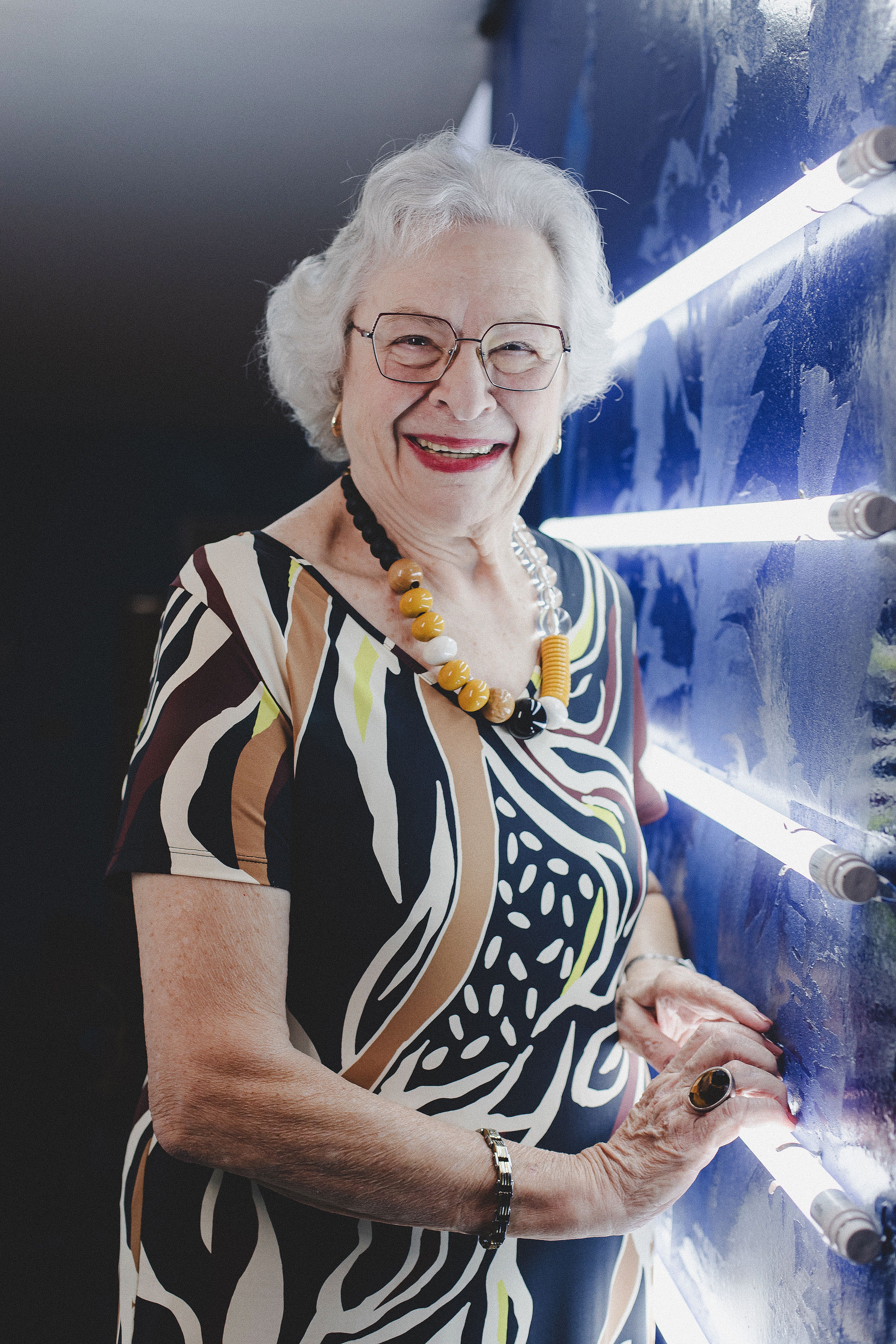A stylish senior woman with gray hair and a bold dress posing by neon lights indoors.