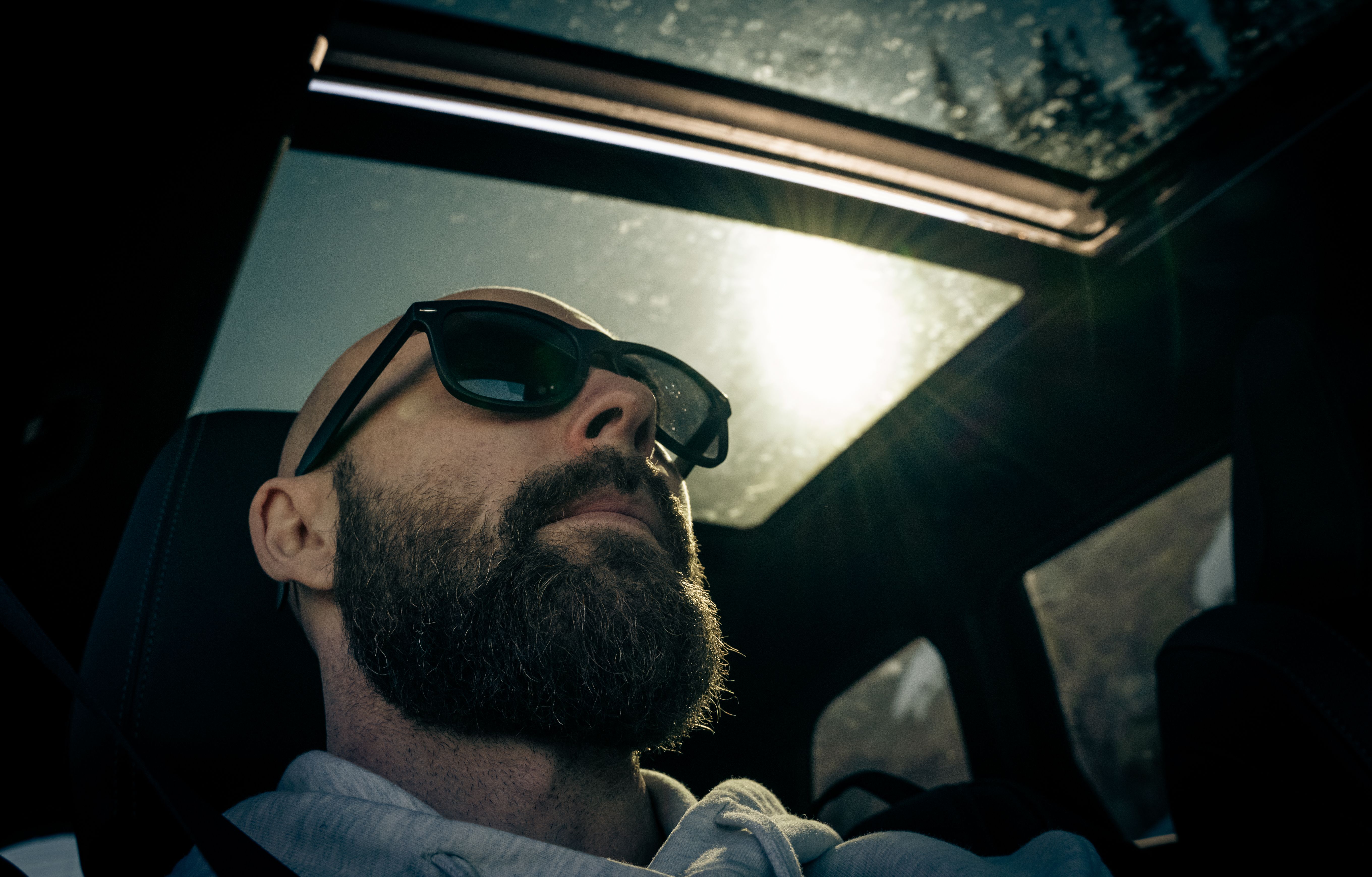A man wearing sunglasses inside a car, enjoying sunlight through the sunroof.