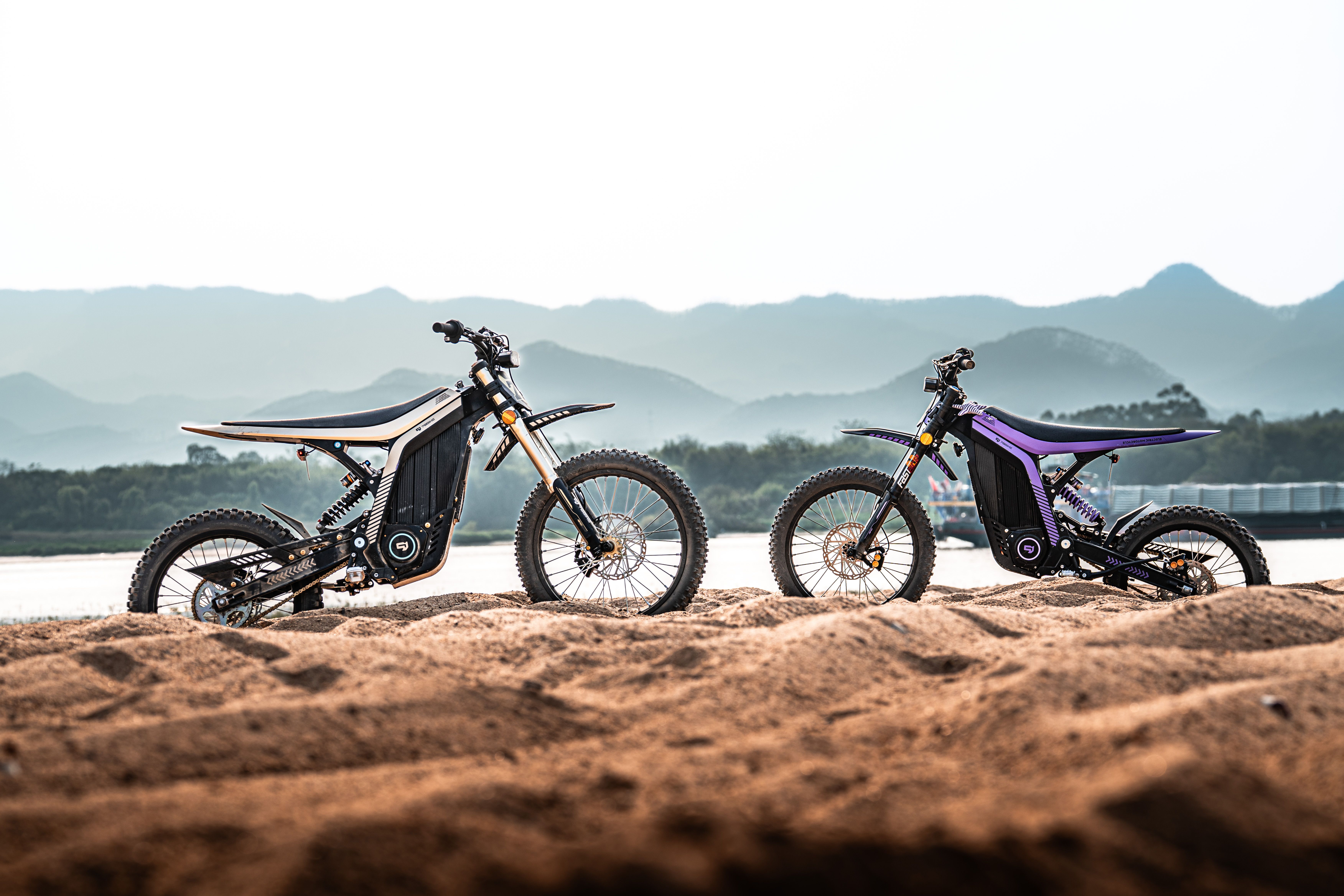 Two electric dirt bikes on sandy terrain against a mountain backdrop at sunrise.