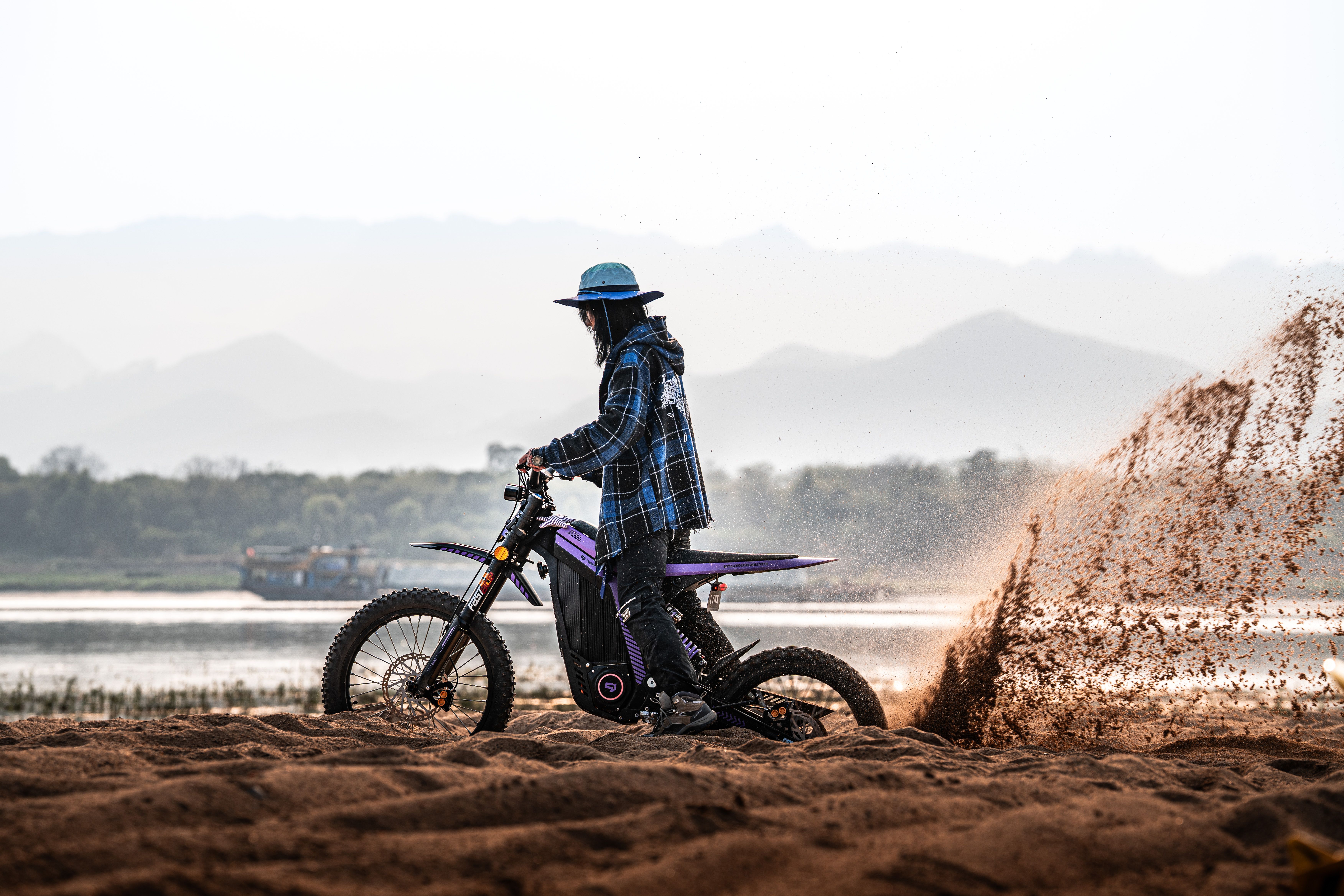 Motorcyclist riding on sand beside a river with mountains in the distance.