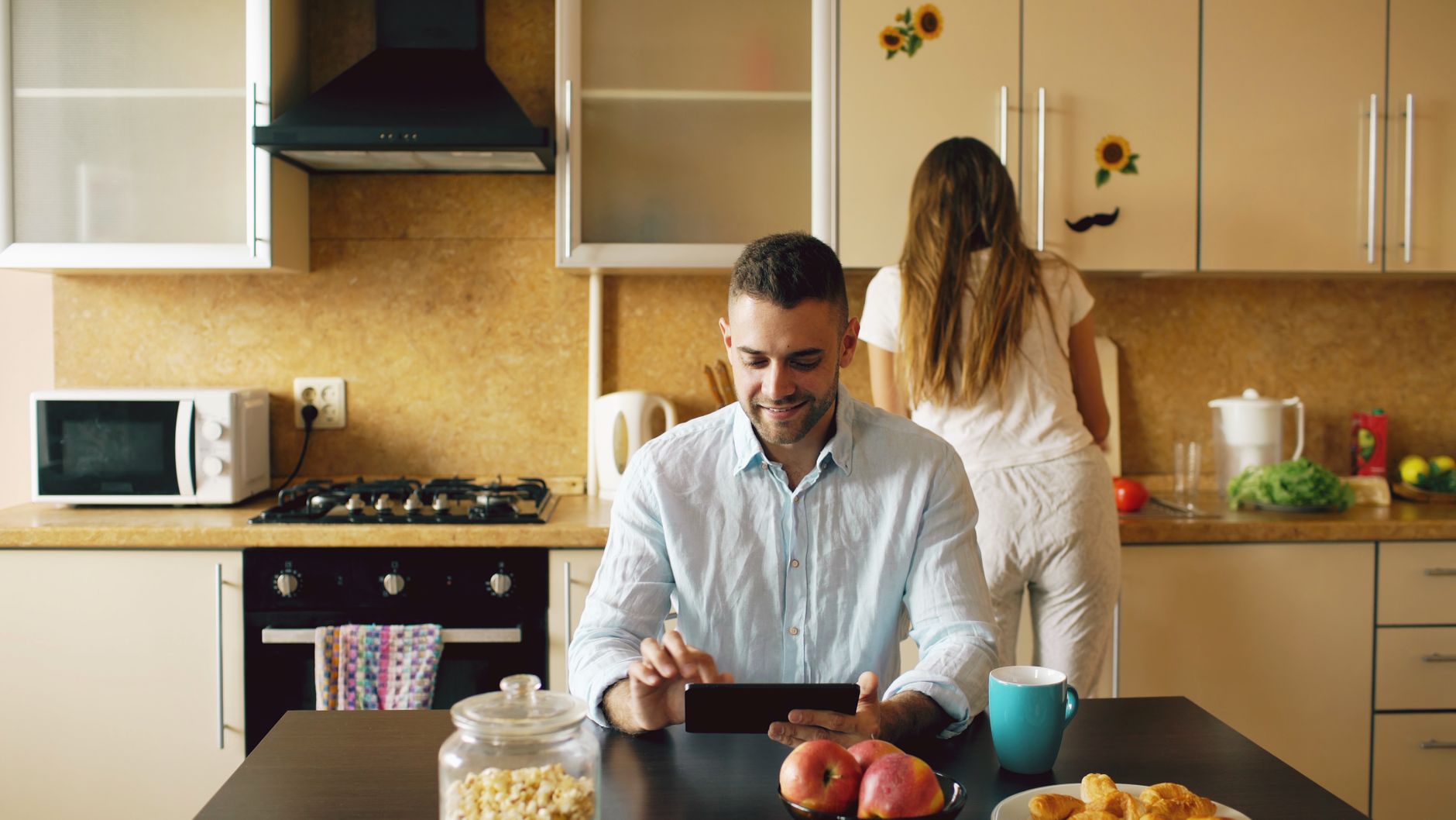 A man using a tablet while sitting at the kitchen table, with a woman preparing breakfast.