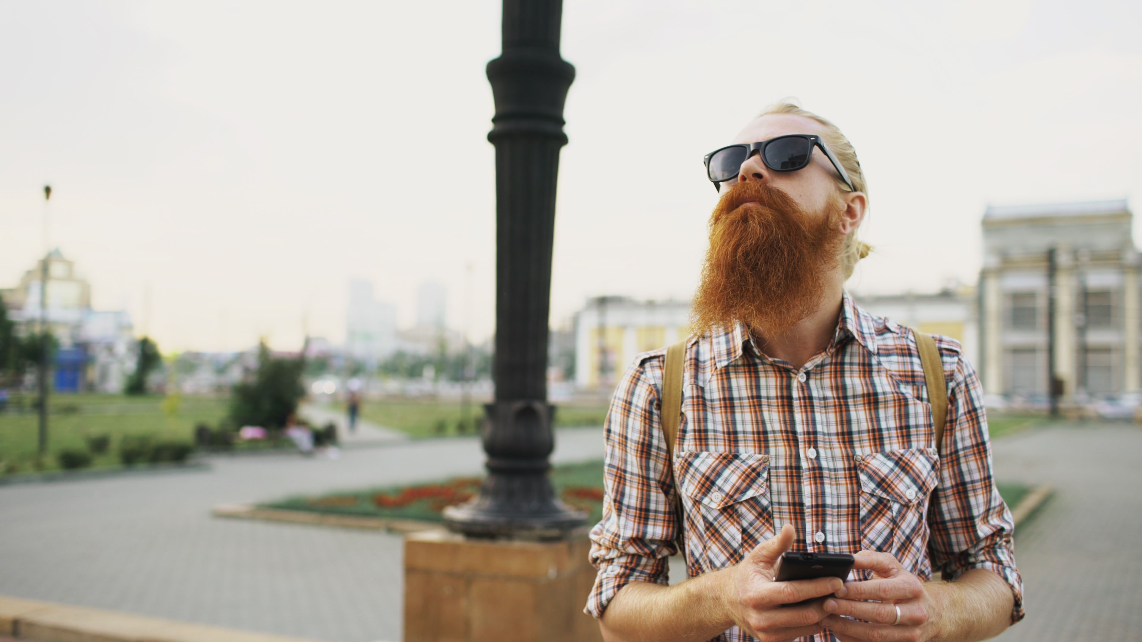 Stylish bearded man in sunglasses using smartphone, outdoors with urban background.