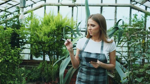 Free Woman in checkered apron observing plants in a greenhouse setting. Stock Photo