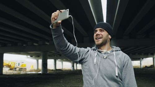 Free Smiling man wearing a beanie takes a selfie outdoors. Urban backdrop. Stock Photo