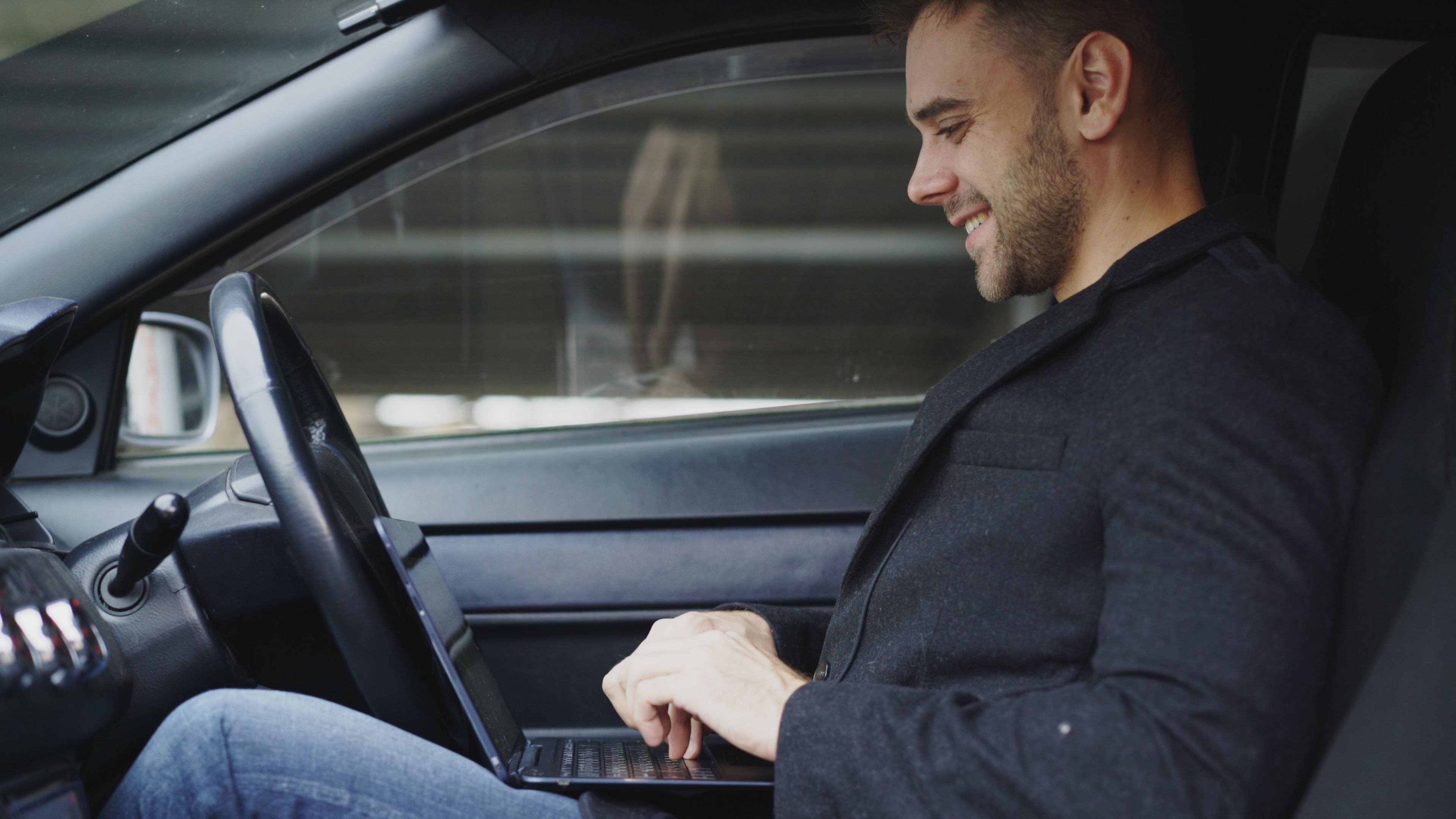 A man in casual attire is smiling while working on his laptop inside a car.