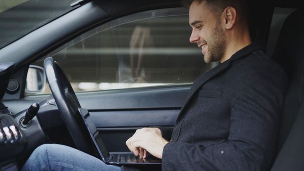 A man in casual attire is smiling while working on his laptop inside a car.