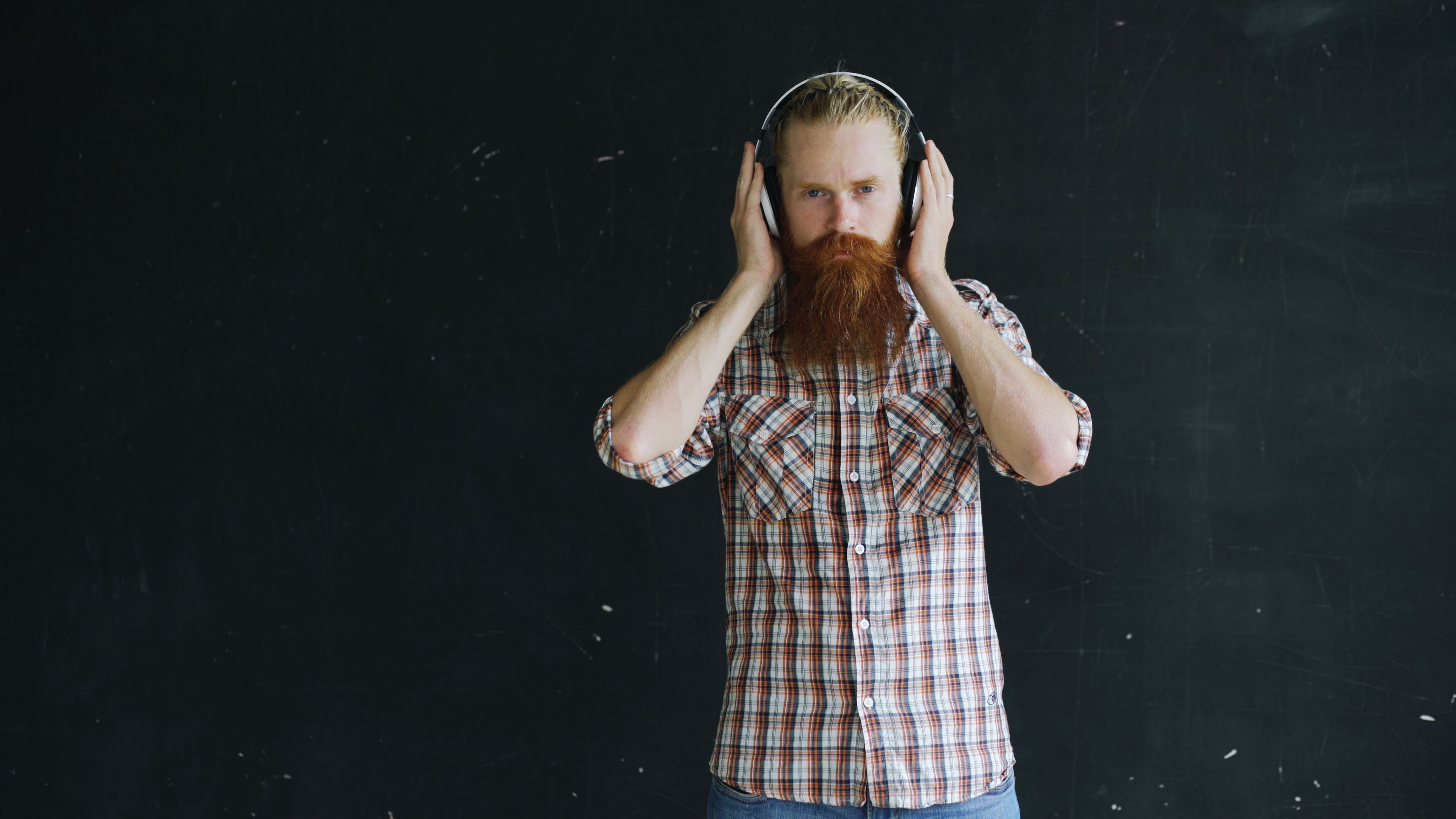 A hipster man with a long red beard wearing headphones against a chalkboard background.