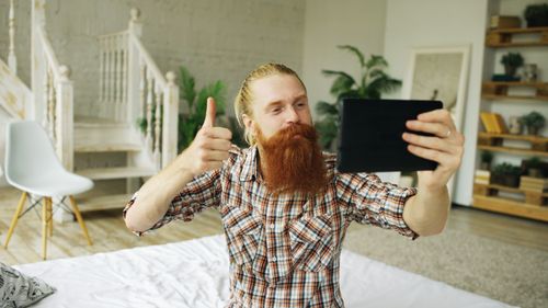 Free Red-bearded man gives thumbs-up while taking a selfie with tablet indoors. Stock Photo