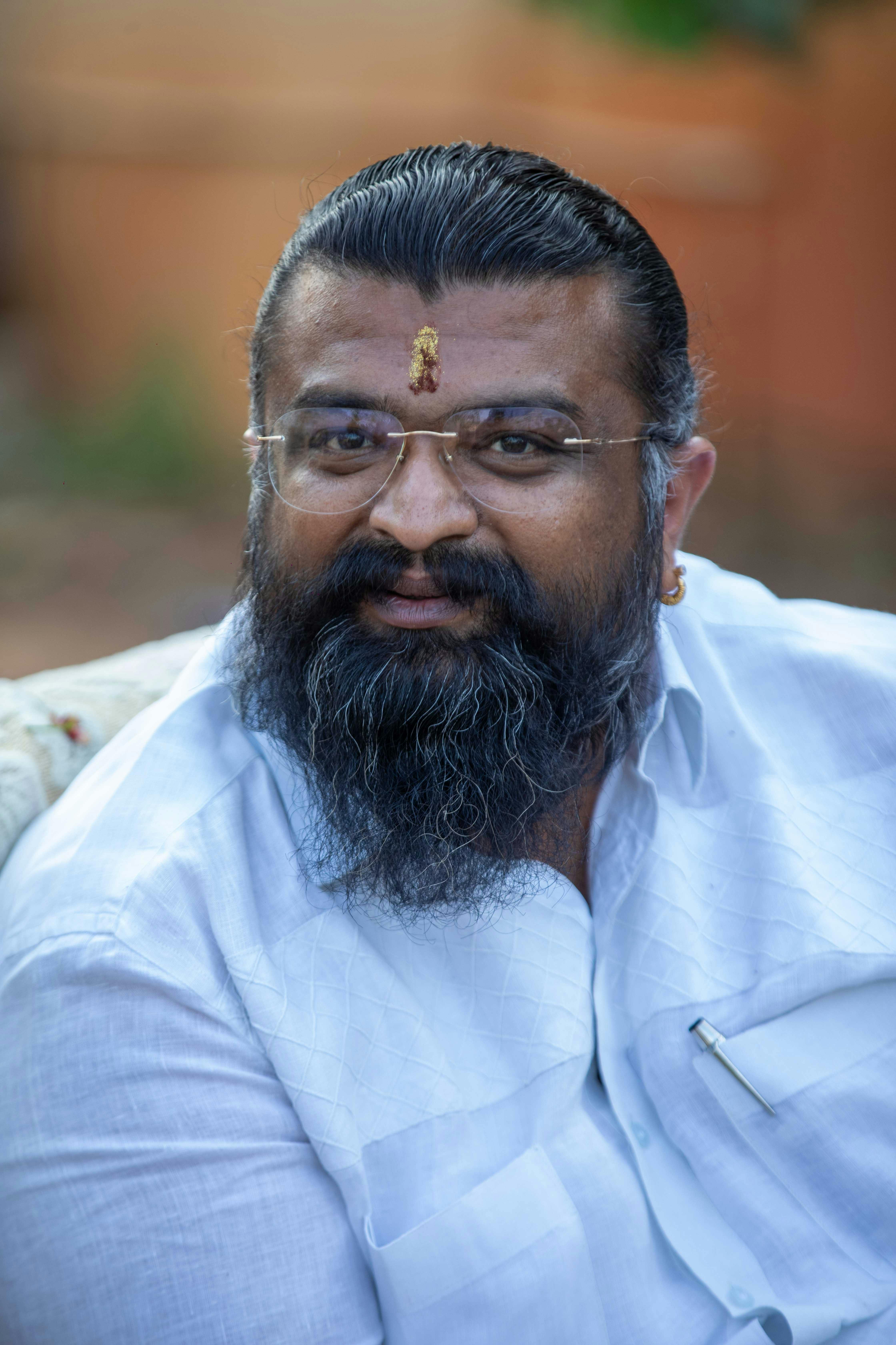 A bearded man wearing glasses and white traditional attire smiles outdoors.