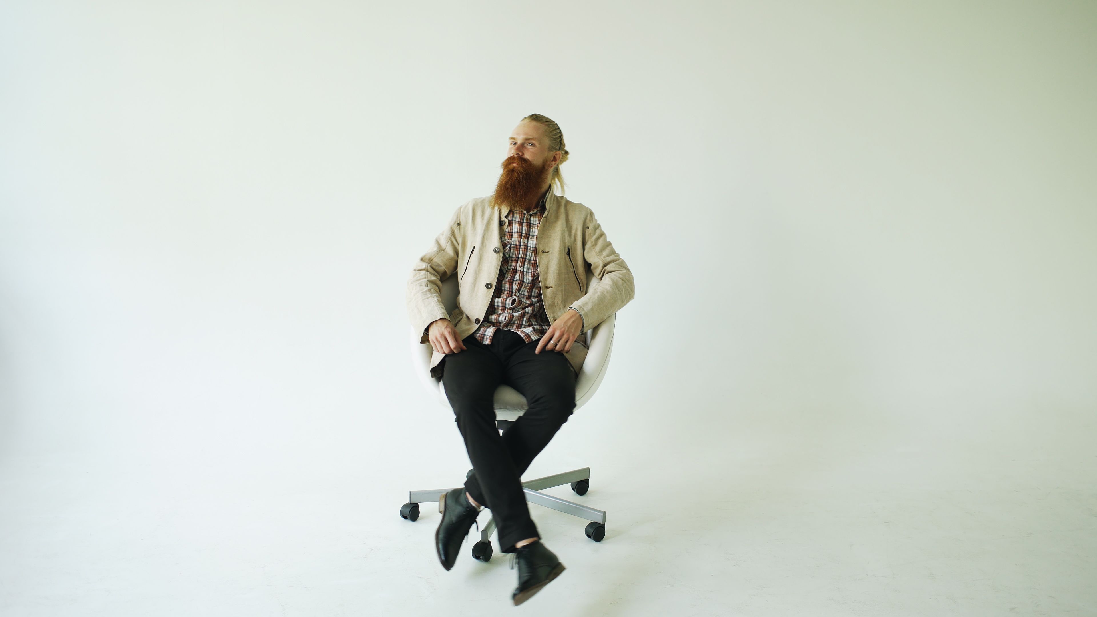 Bearded man in beige jacket sitting on a white swivel chair in a minimalist studio setting.