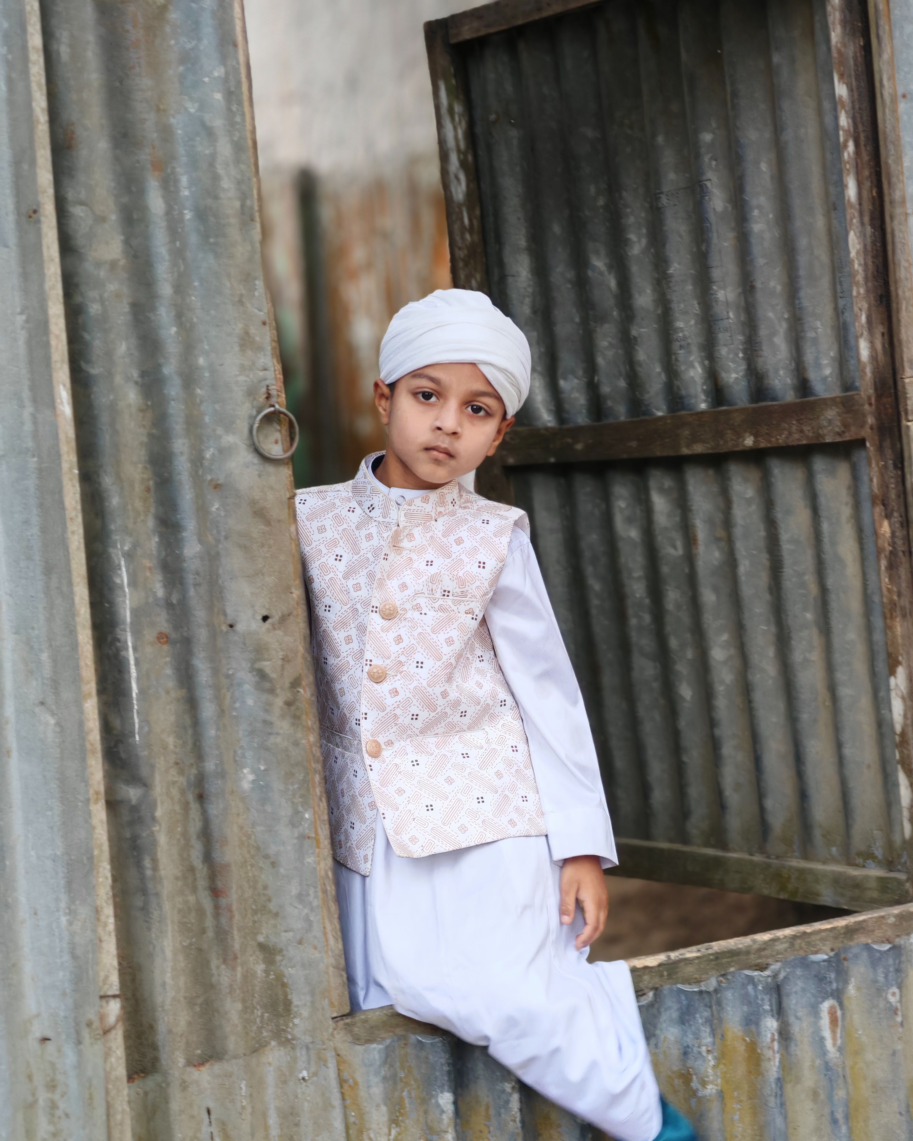 A young boy wearing traditional clothing poses by a rusty corrugated metal gate, showcasing cultural fashion.