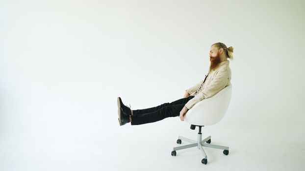 A man with a beard relaxes in a white chair, in a minimalist, bright setting.