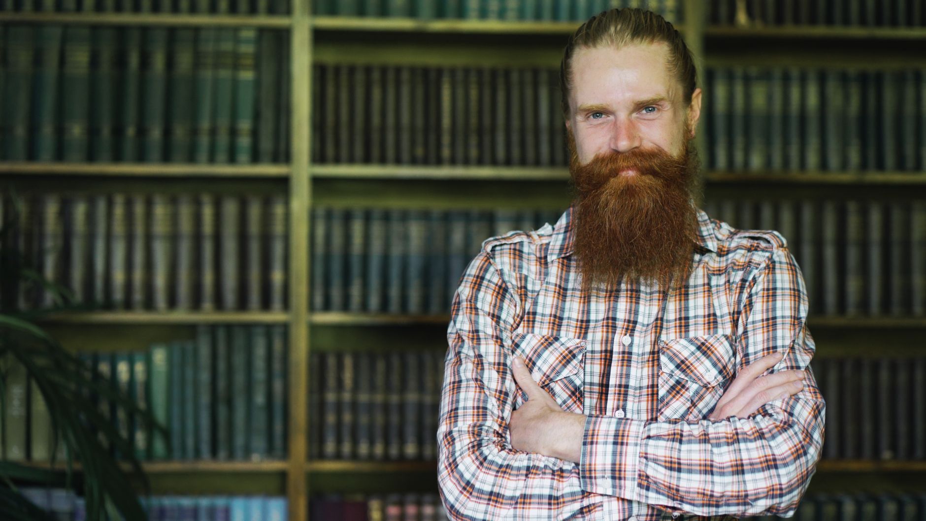 Man with a long beard standing confidently in front of bookshelves in a library.