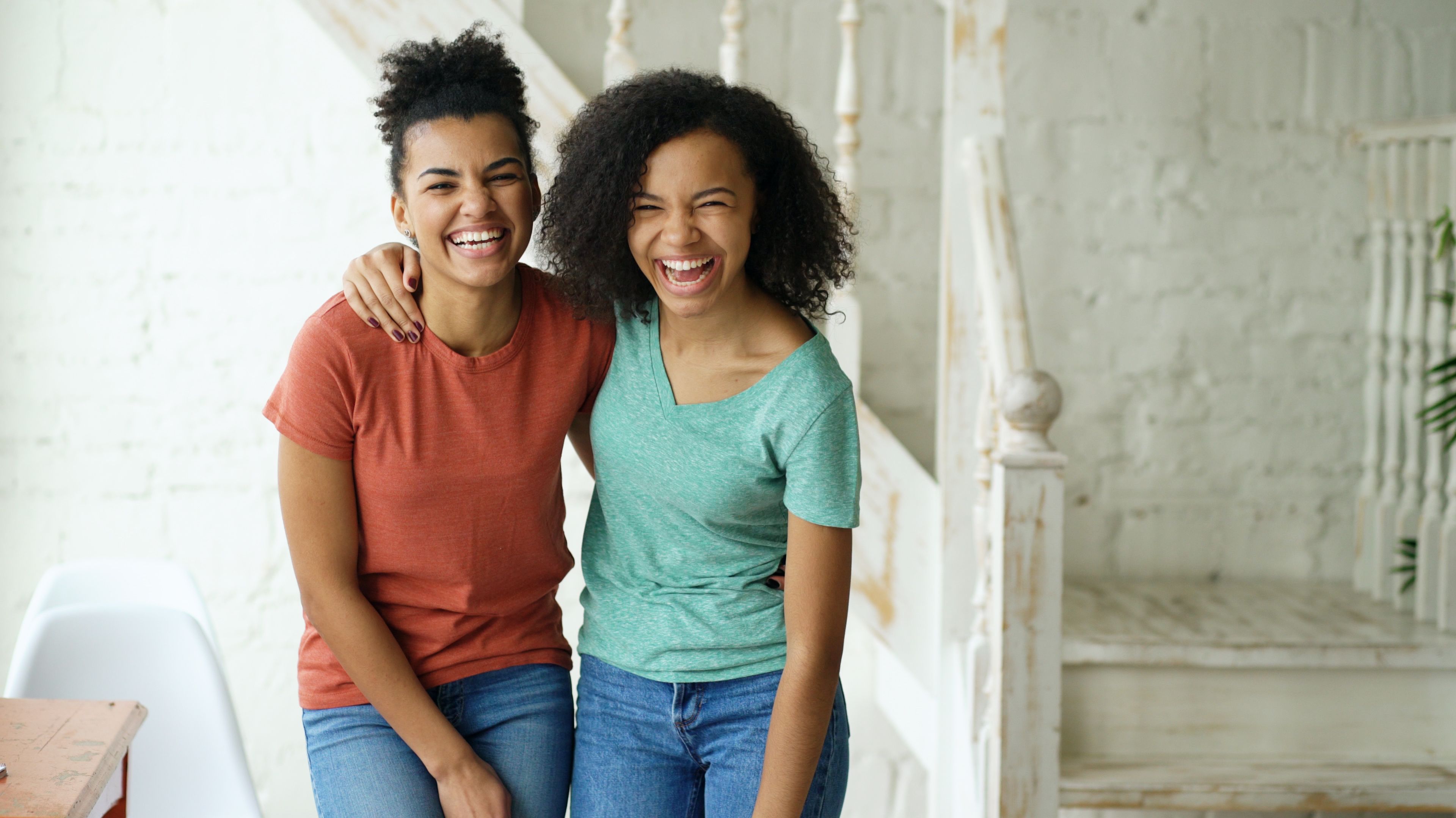 Two women in colorful shirts laughing happily indoors, embracing friendship.