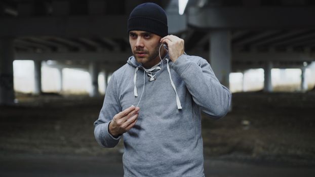 Athletic man under bridge adjusting earphones for run, capturing urban fitness vibe.