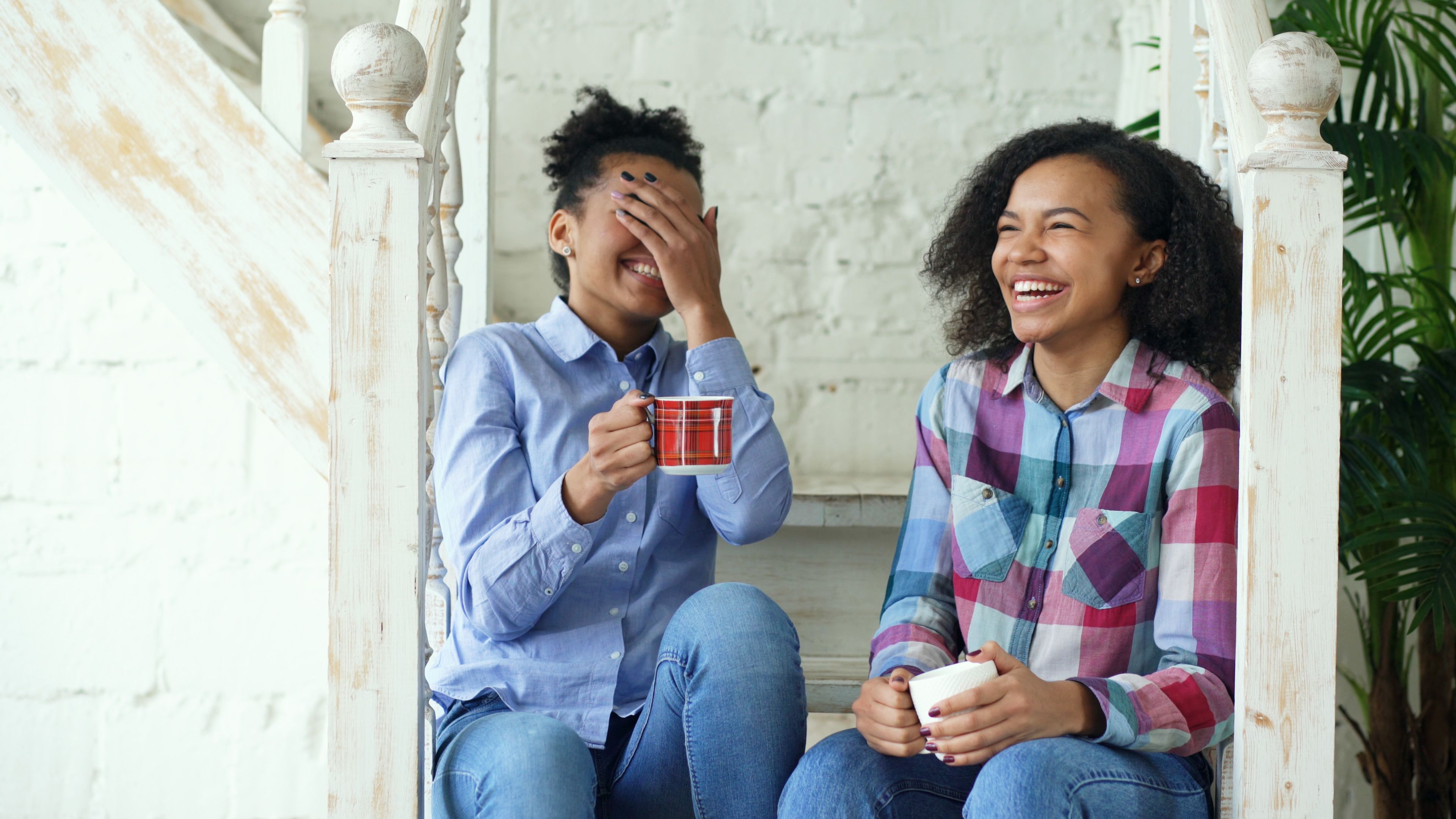 Two smiling women enjoy coffee on stairs, sharing laughter and friendship indoors.