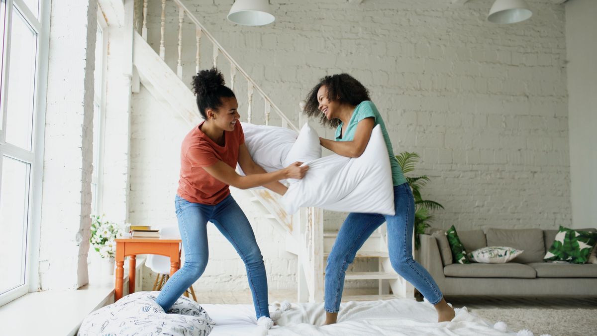 Two women having a playful pillow fight in a sunlit modern living room.