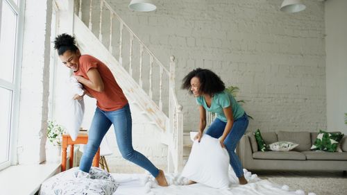Free Two women joyfully engage in a playful pillow fight indoors, laughing and having fun. Stock Photo