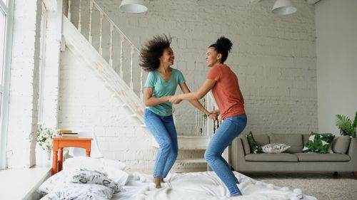 Free Two women joyfully jump on a bed, creating a lively and playful scene indoors. Stock Photo