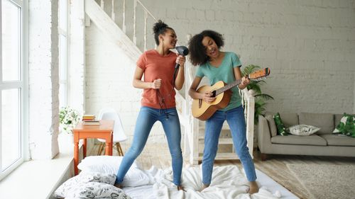 Free Two women having fun singing with a hairdryer and guitar on a bed in a cozy living room. Stock Photo