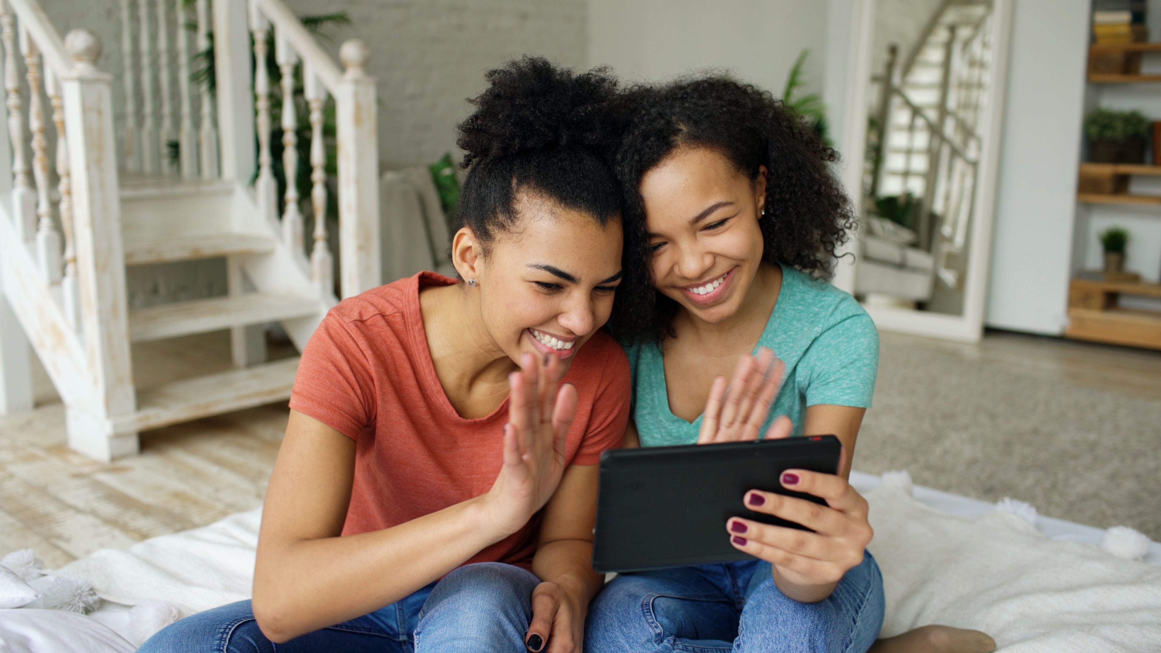 Two friends enjoying a video call on a tablet at home, smiling and waving.