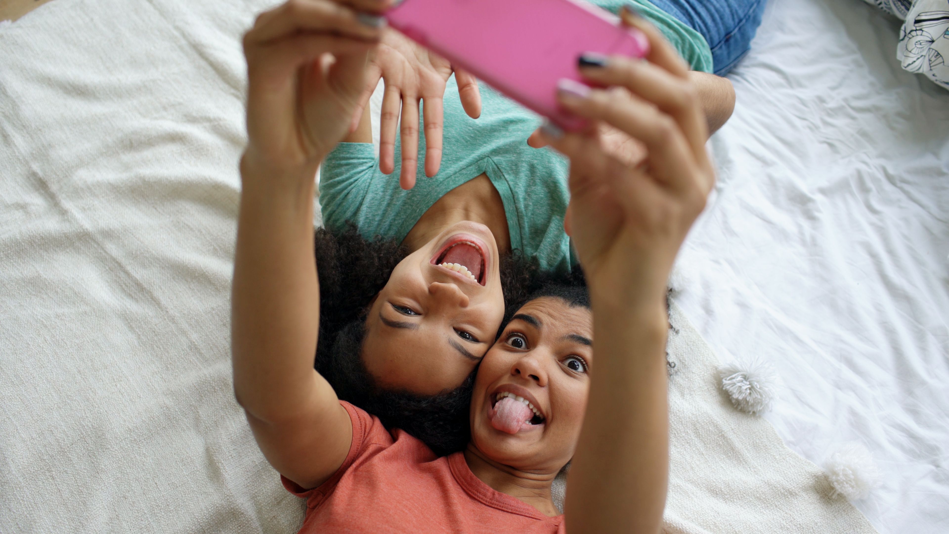 Two smiling teenagers lying on bed taking a playful selfie with smartphone.