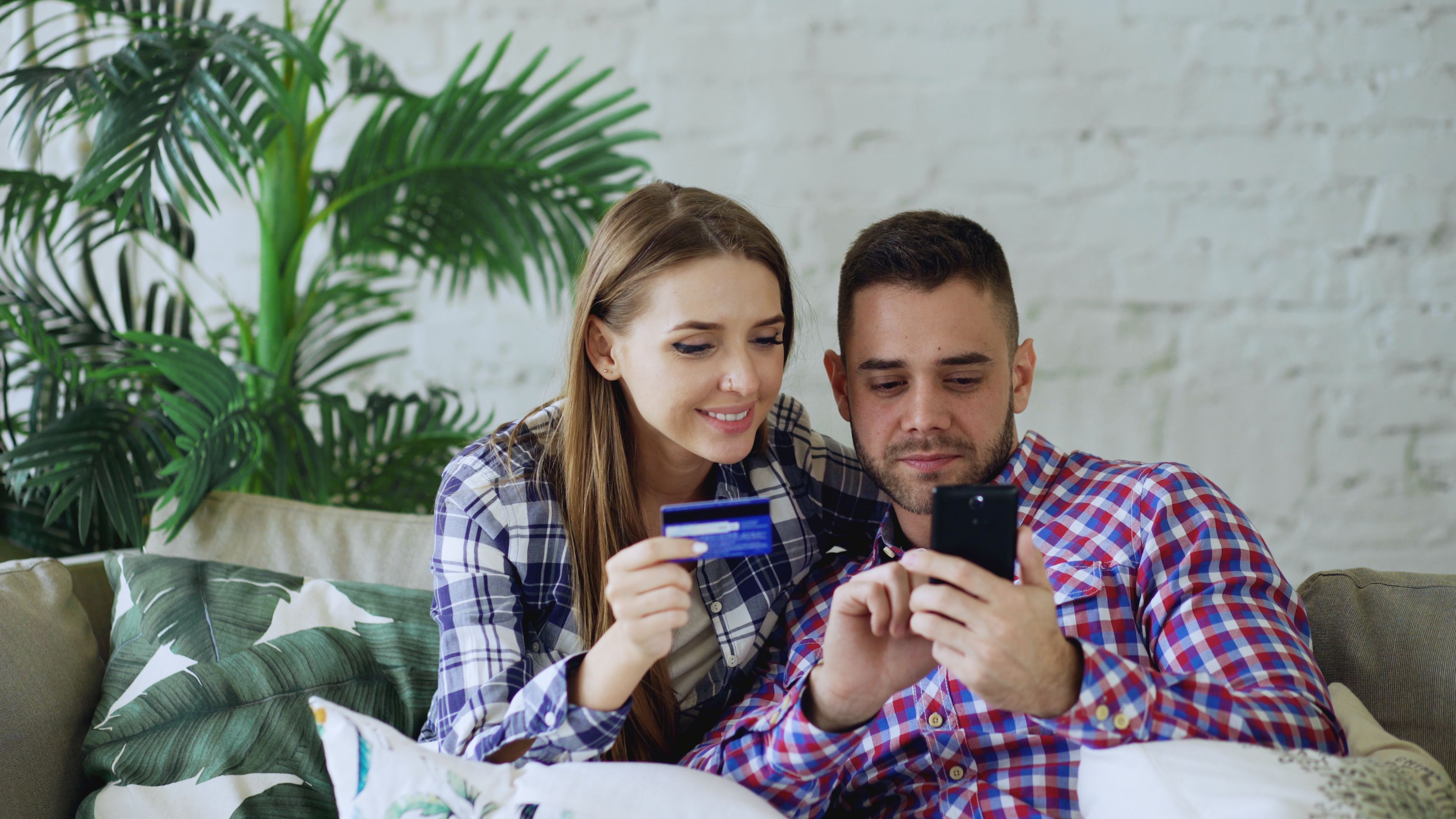 Smiling couple sitting on a sofa, shopping online with a smartphone and credit card.
