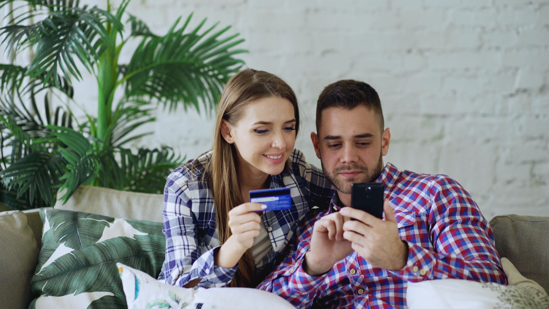 Smiling couple sitting on a sofa, shopping online with a smartphone and credit card.