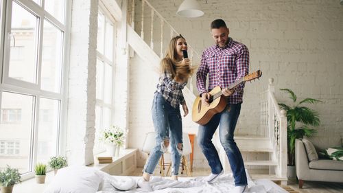 Free Joyful couple singing and playing guitar on a bed in a bright, cozy room. Stock Photo
