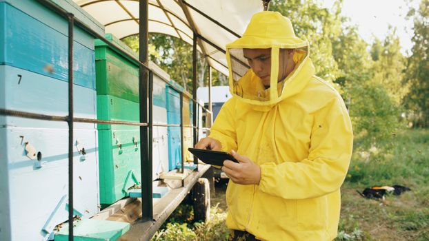 Beekeeper wearing yellow suit inspects beehives in apiary on a sunny day outdoors.
