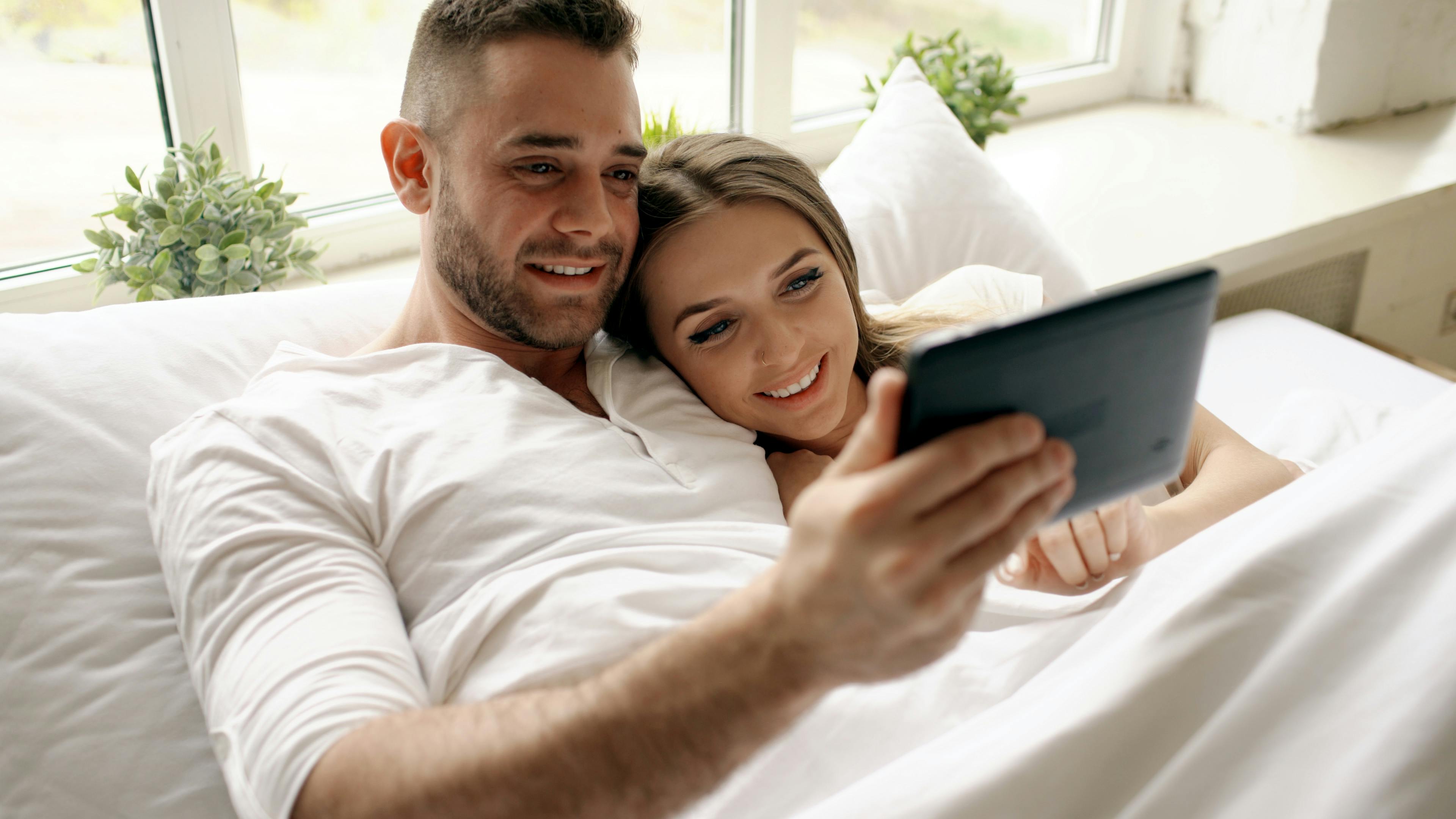 Two people holding a tablet and cuddling in bed