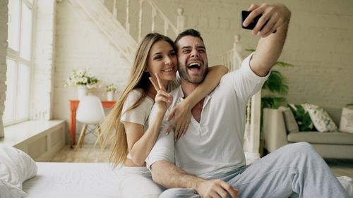 Free Cheerful couple taking a selfie in a cozy, modern bedroom setting. Stock Photo