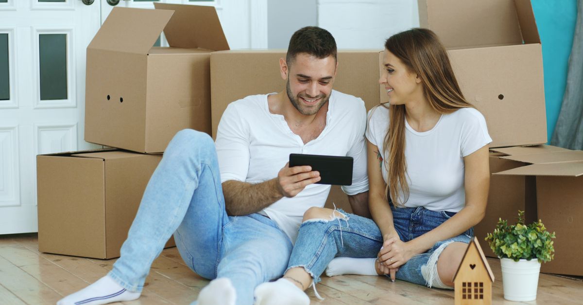 Young couple happily unboxing in their new home, viewing something on a tablet.