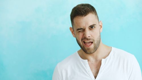 Free A young man playfully winks with a cheerful expression against a blue background. Stock Photo