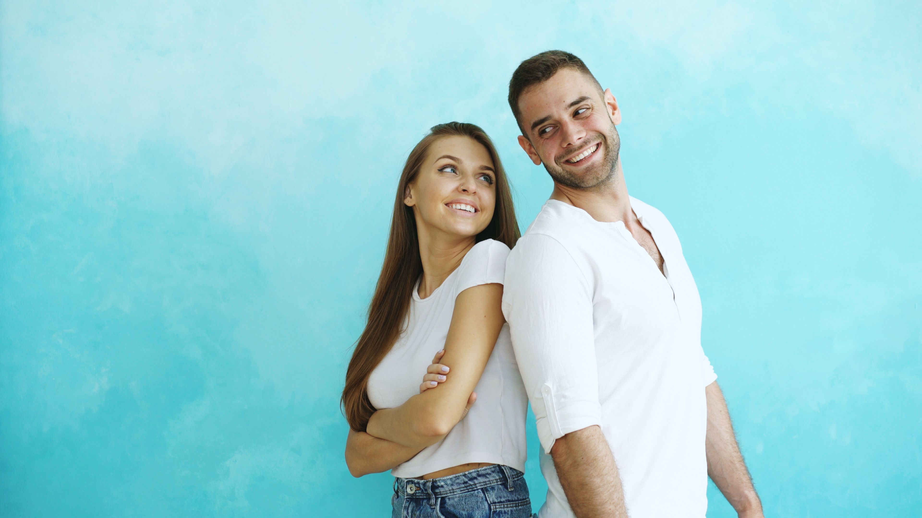 Happy couple in white shirts pose playfully against a bright turquoise backdrop.