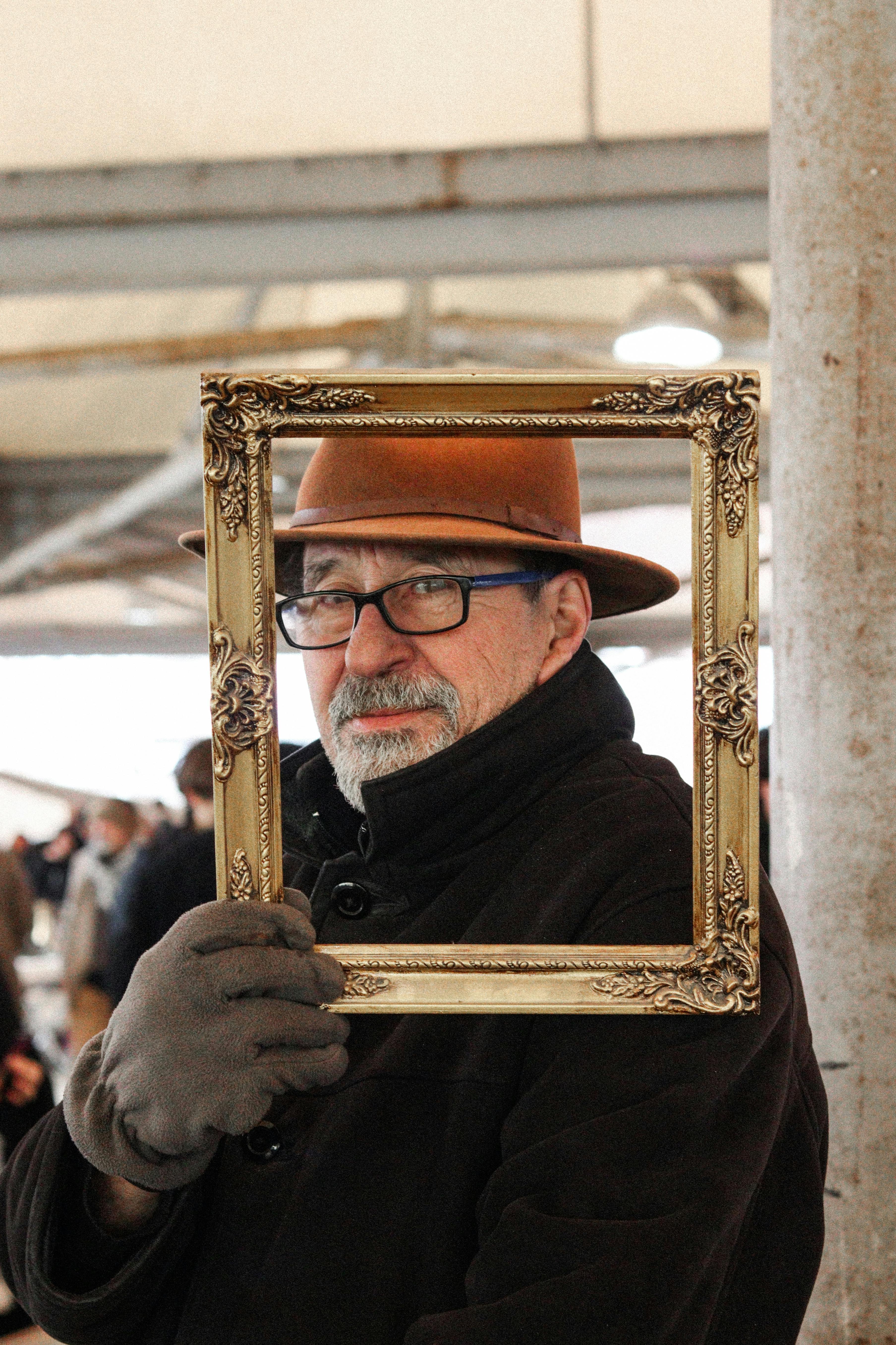 Elderly man with glasses and hat framed in gold antique frame indoors.